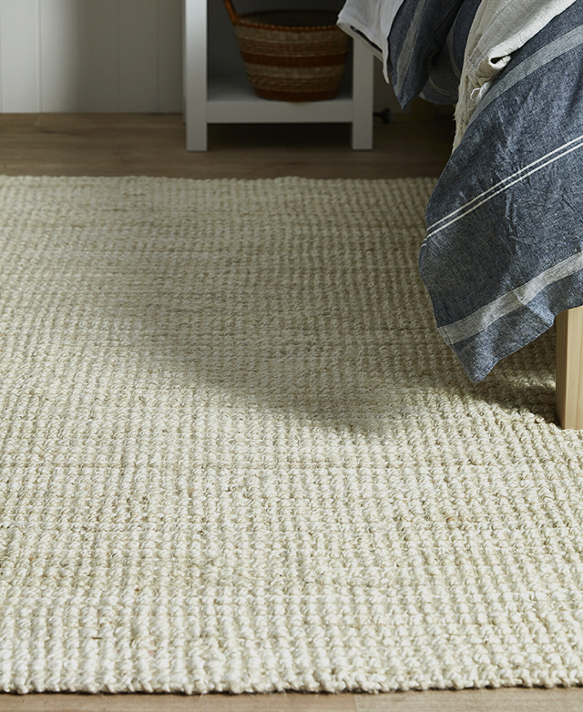 The side view of the rug brings out the weave detail of the soft, neutral floor decor, which is positioned next to a bed.