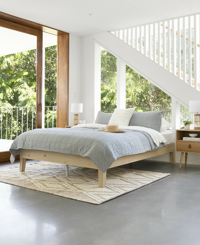 A fully made bed in a light-filled room, with the bed's front legs standing atop a textured rug.