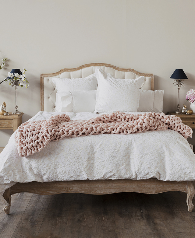 The bed is made with white sheets, with a pink knitted blanket draped over the middle. Side tables & lamps frame the bed.