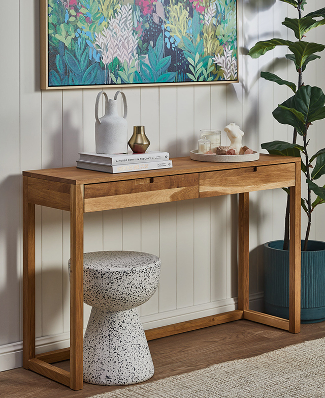 Solid oak table with decor on top is positioned underneath a colourful botanical artwork and next to a tall fiddle leaf fig.