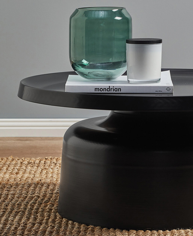 The coffee table is pictured from the side where the curved pedestal is centre stage. A book, candle & vase are on top. The coffee table is pictured from the side where the curved pedestal is centre stage. A book, candle & vase are on top.