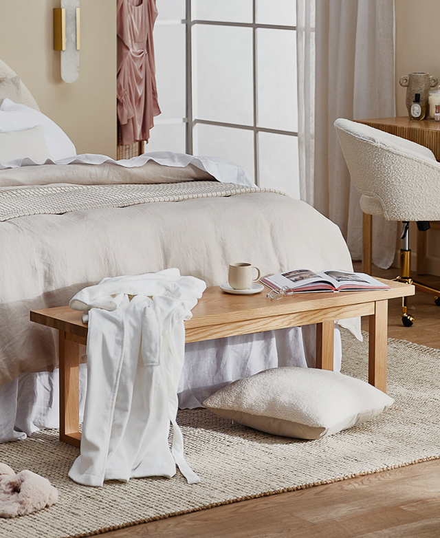 A bench positioned at the foot of a bed; on top, a book is open next to a coffee mug and glasses, and a robe is draped over.