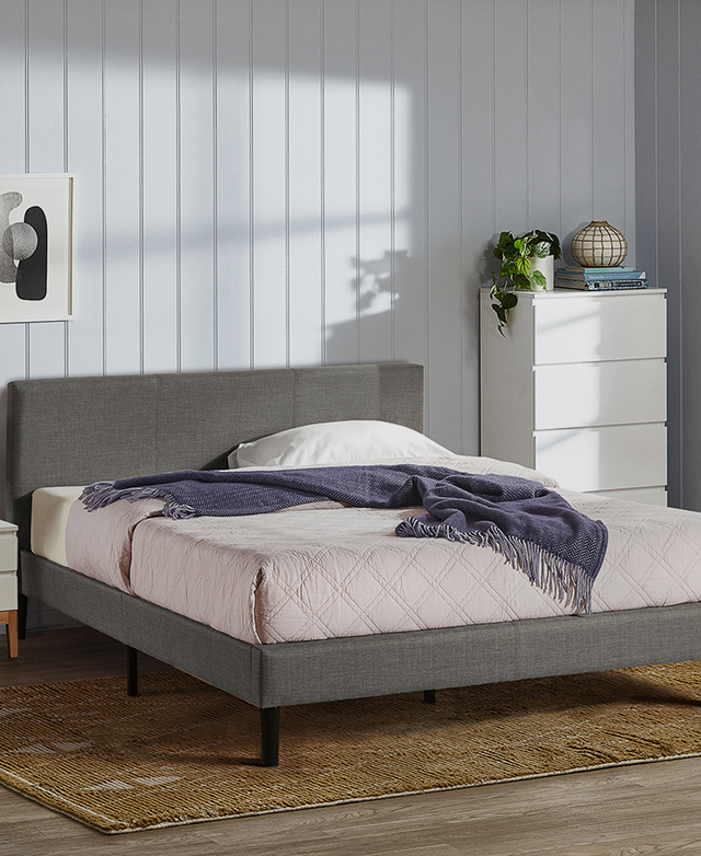 Soft blue panelled walls in a gently lit room contains the assembled and dressed bed. A golden rug is under the bed.