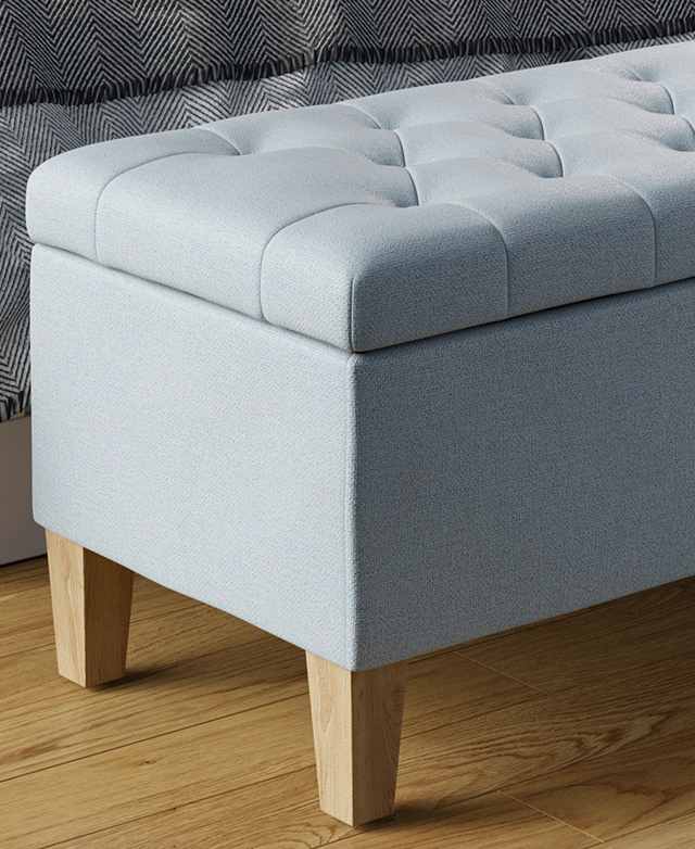 On top of timber flooring, an ottoman with baby blue fabric upholstery is positioned at the foot of a bed.