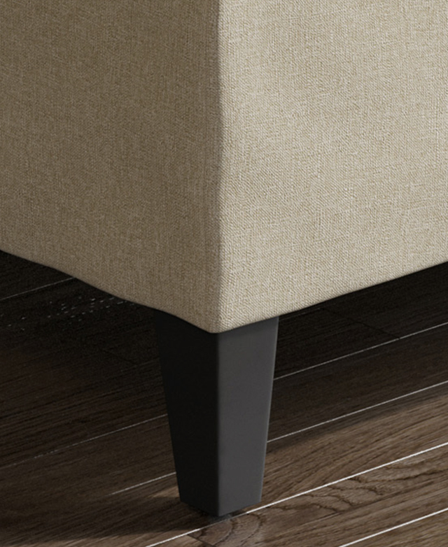 Large-scale view of a rubberwood leg underneath the beige ottoman. It has a black finish and chunky tapered shape.
