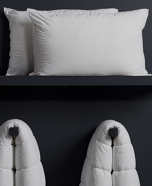 Against a black textured wall, two plump pillows are stored on a high shelf. Two quilts hang on hooks below. Against a black textured wall, two plump pillows are stored on a high shelf. Two quilts hang on hooks below.