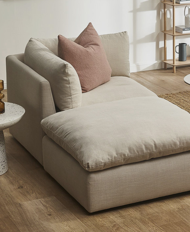 In cream upholstery, a corner modular sofa chair and large ottoman are pushed together, forming a chaise lounge.
