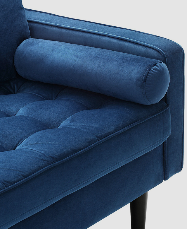 One end of a sofa with blue velvet upholstery is shown. A plump bolster cushion rests against the low rectangular armrest.