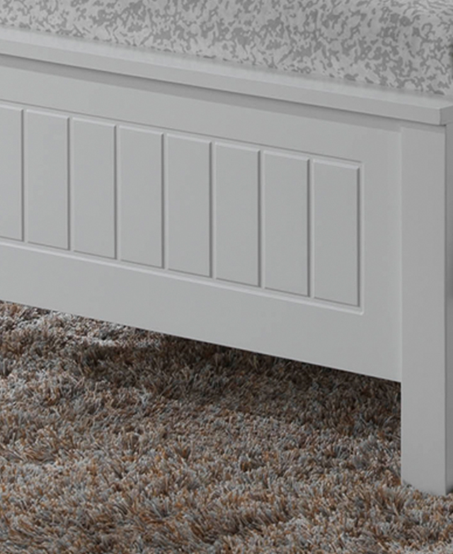 The end of a bed frame atop a shaggy rug. It has solid rubberwood legs, and panel details etched into the white footboard.