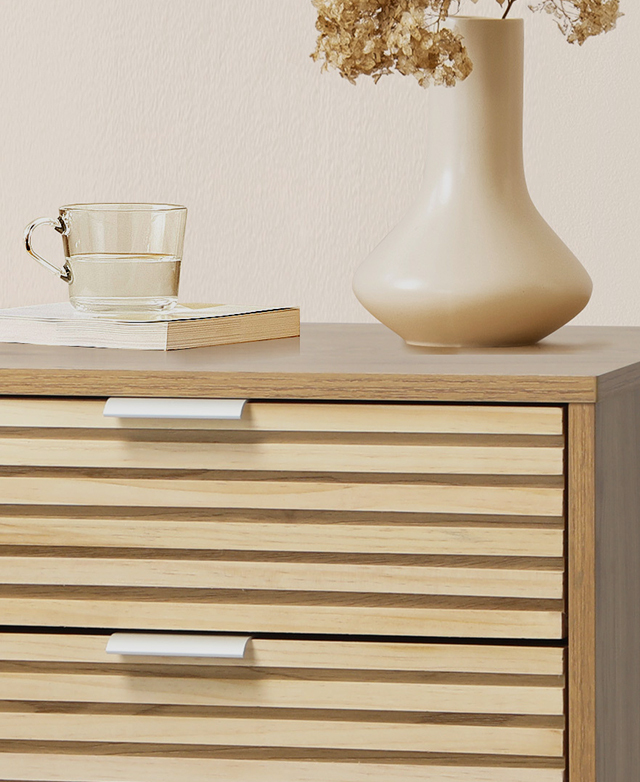 The horizontal panelling detail on the face of both drawers is in focus. A book, tea cup, and vase of dried florals sit on top of the bedside table. The horizontal panelling detail on the face of both drawers is in focus. A book, tea cup, and vase of dried florals sit on top of the bedside table.