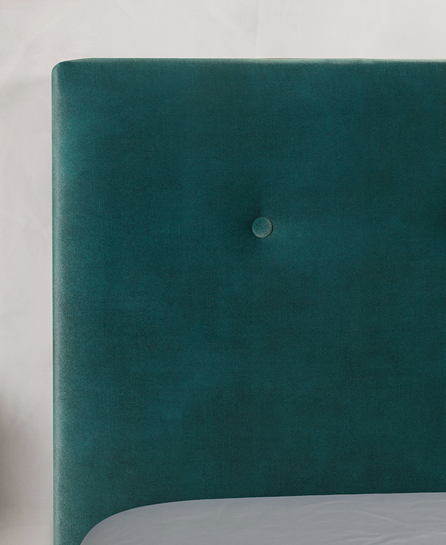 A corner of the bedhead is in focus, highlighting the emerald green velvet upholstery and button-tuft design detail.