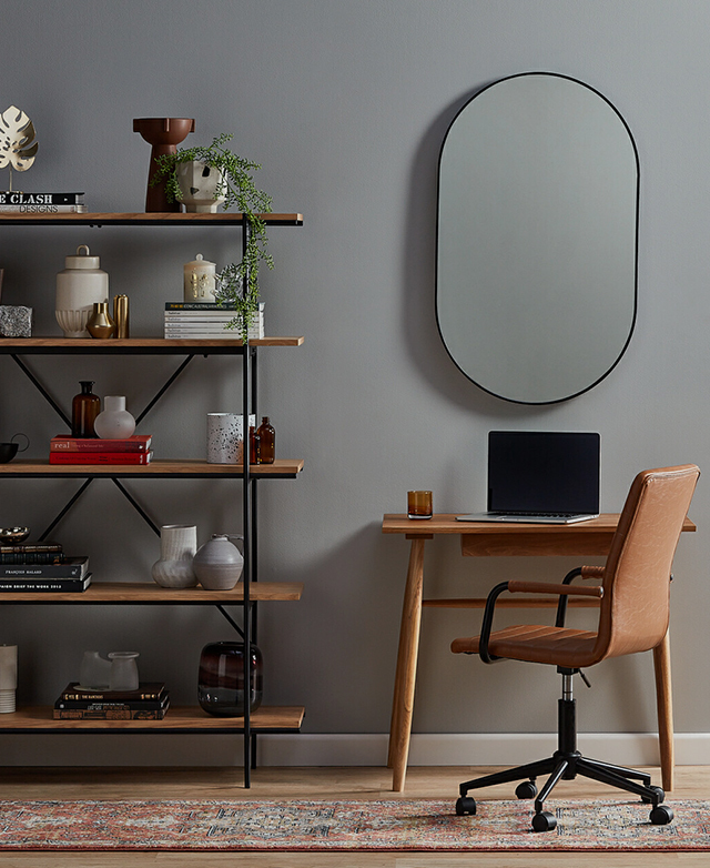 An oval mirror with a black frame styled above a small wooden desk in a study nook, with an industrial-style bookcase next to it.