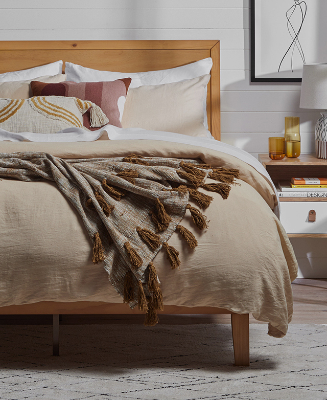 A bed with a timber bed frame is dressed in beige and white sheets, with a fringed throw draped over the top.