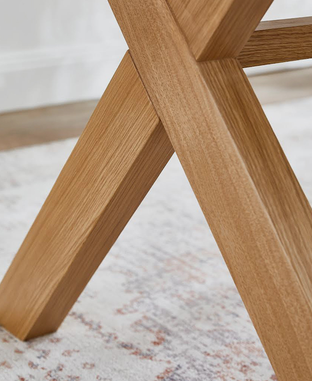 The X-shape of the timber dining table frame is in focus. It's styled above a rectangular area rug. The X-shape of the timber dining table frame is in focus. It's styled above a rectangular area rug.
