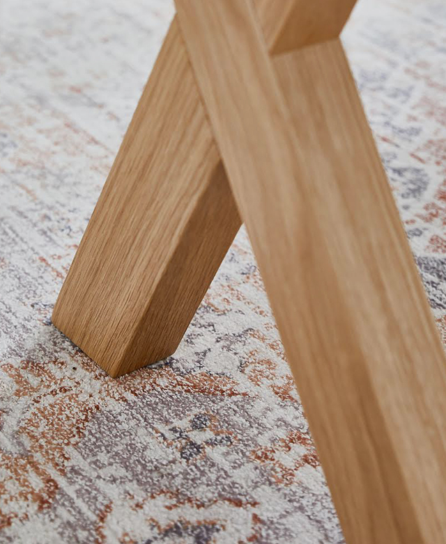 The X-shape base of the timber bench is in focus. It's styled on top of a patterned, low-pile rug.