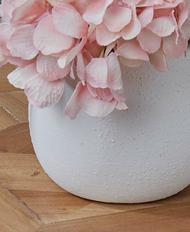 Soft pink petals of a faux hydrangea flower cascade down a textured white ceramic pot.