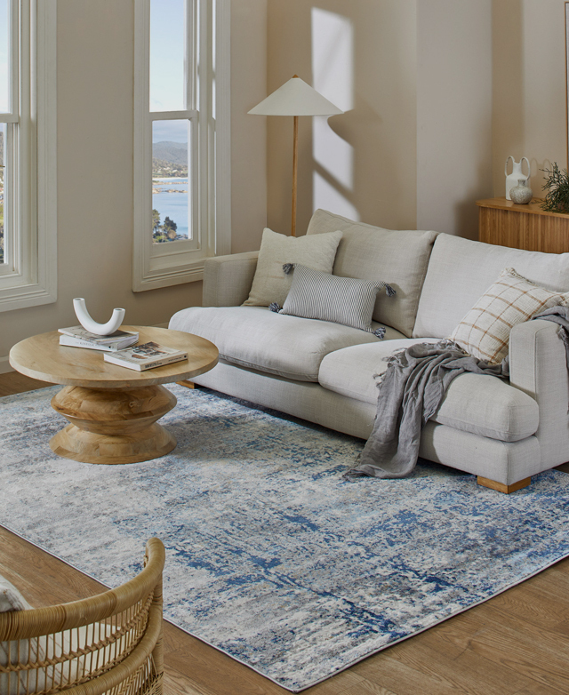 In a bright room, the rug is positioned with a grey armchair and ottoman, with a blue cushion and ceramic respectively.