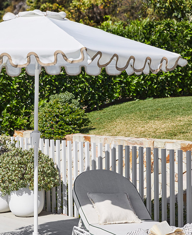 The sage-coloured scalloped umbrella is erected in an outdoor setting, near grass, a white picket fence, and a sun lounger.