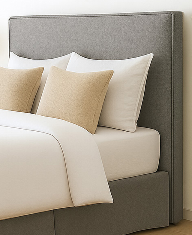 Three-quarter angle of a bed and its grey piped bedhead, with cushions arranged in front of it.