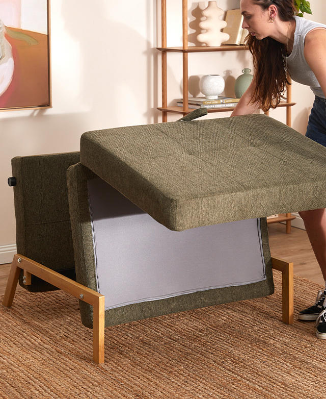 The green sofa bed is being unfolded by a woman on a taupe rug.