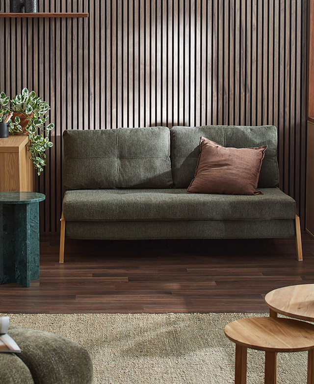 A front view of the green sofa bed is positioned in the corner of a walnut-clad room.