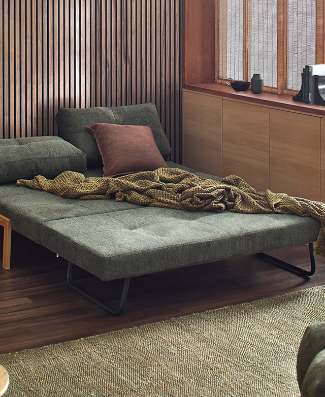 The green sofa bed is unfolded into bed formation, and has a rust-coloured cushion and green blanket on top.