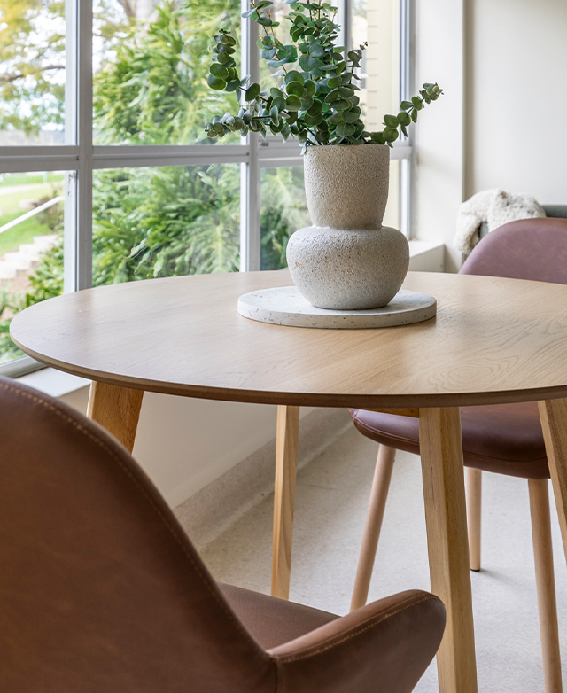 Cropped view of the 'natural' dining table is positioned in front of a panelled window with leather dining chairs.