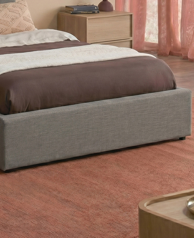 The end of the grey upholstered bed with mattress is atop a terracotta rug.