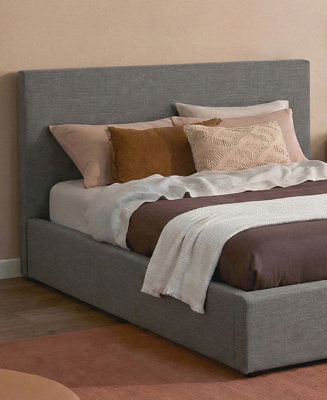 The grey upholstered bed with bedhead is in front of a peach wall and topped with several pillows and cushions.