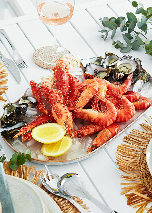 A long platter sitting on a table filled with cooked prawns, oysters and slices of lemon