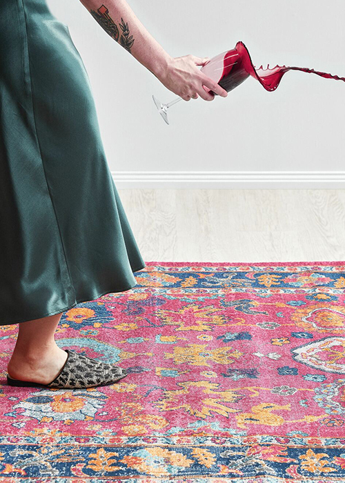 A person stepping onto a colourful rug, holding a glass of red wine that is spilling over the edge of the glass and onto the rug