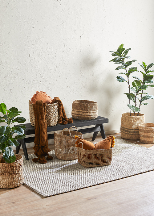 Seven woven baskets arranged around a black bench some with plants in them and some with muted orange blankets and cushions in them