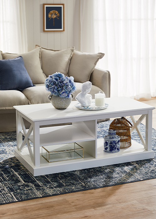 A Hamptons style living room focused on the coffee table which has a small tray with a bust sculpture and candle on it as well as the sofa which has three beige cushions and one navy cushion