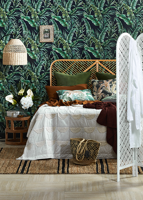An island inspired bedroom with palm leaf wallpaper and a woven white room divider with a bathrobe hanging off it in front of the bed