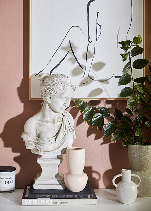A close up shot of a marble bust of a woman with a flowing toga and wavy hair on top of a stack of two books next to two small vases and a leafy plant