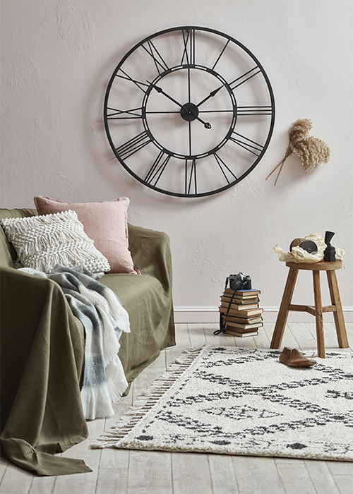 A living room with a olive coloured sheet over the sofa a stack of nine A5 books with a camer a on top and a large black metal industrial style clock on the wall with roman numerals around the face