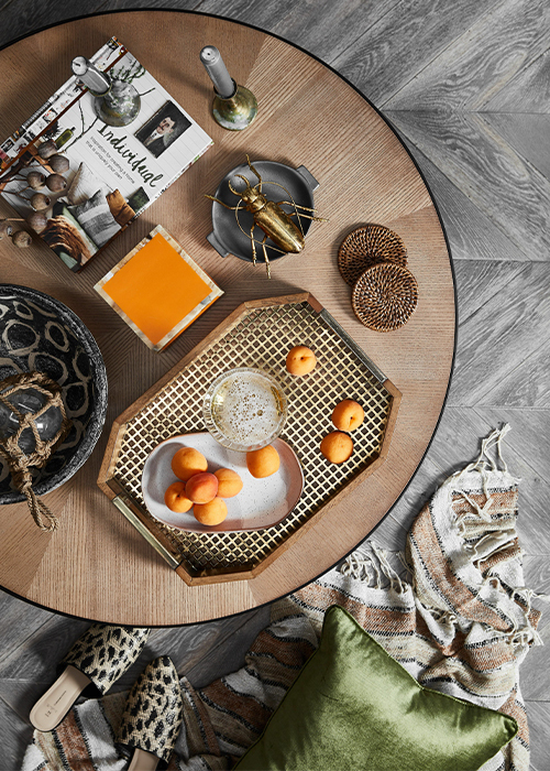 A birds eye view of a coffee table with an ovular ceramic dish of scattered nectarines on a wooden tray with black and gold grid detailing and two decorative bowls, one which has a golden beetle ornament in it