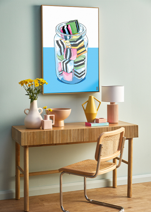 A light wood desk with fluted detailing and a Cesca cane chair with three pastel coloured sculptural vases a peach coloured bowl and peach coloured lamp on the desk, with a large painting of a jar of liquorice allsorts on the wall