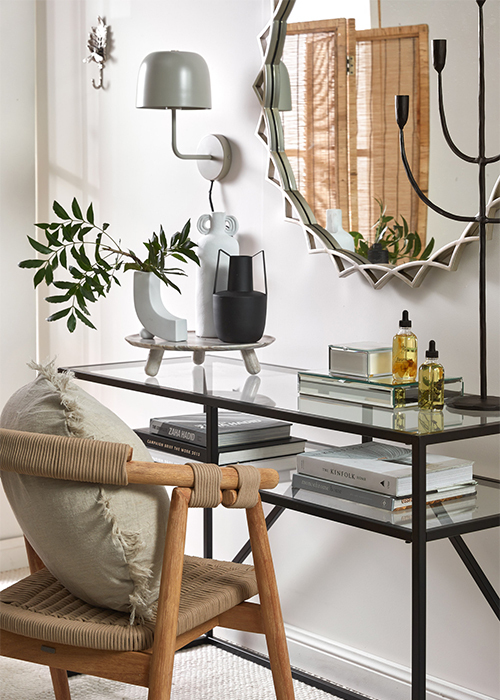 A modern style glass dressing table with an large ornate sun shaped mirror hung on the wall and a rattan accent chair with a beige linen cushion