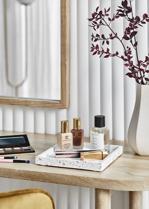 A close up of a square terrazo tray with perfume lipstick and two nail polish bottles on it next to a ceramic vase and eyeshadow palette on a wooden dressing table