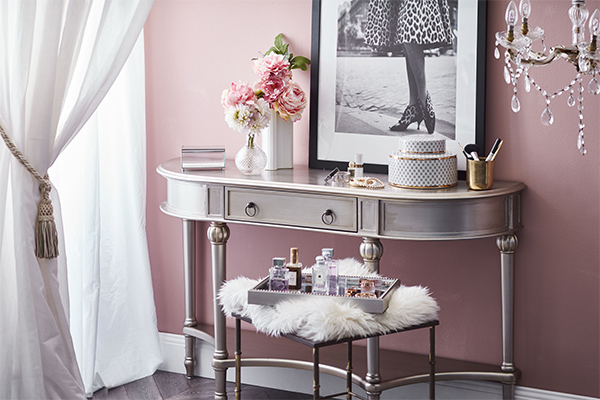 A 1920s style silver dressing table with a black and white artwork of a woman in a leopard print dress leaning against the wall, a jar of makeup brushes and vase of flowers on the table and a faux fur toppede stool with a silver tray filled with perfume bottles on it