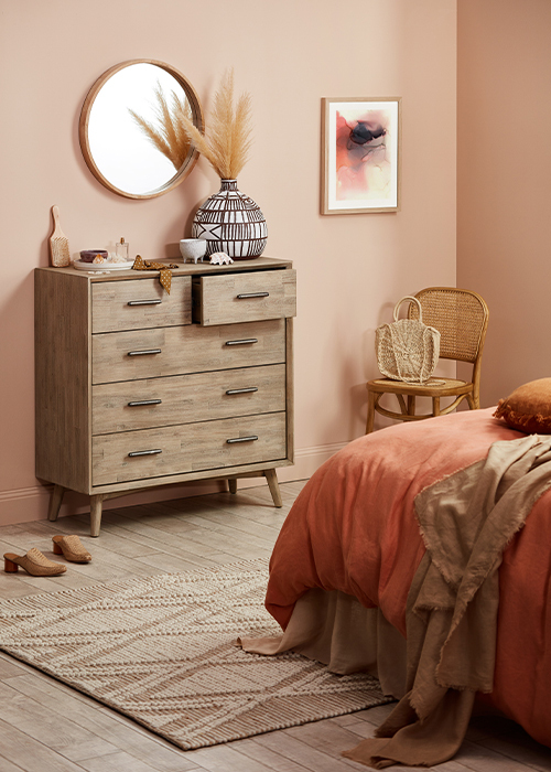 A bedroom with a tall wooden chest of drawers with a large pot holding dried leaves and various personal items like jewellery perfume and a hairbrush on top with a round wooden framed mirror hanging on the wall