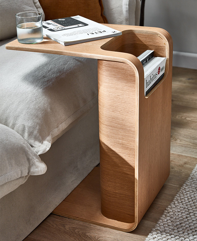 The C-shape fits over a creamy sofa seat, with the top featuring a magazine and a clear glass of water.