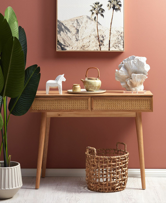 A console table next to a tall plant. Landscape artwork is hung above, and decor items are situated on the top surface.