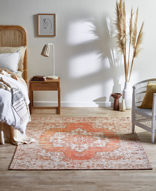 The orange flat weave rug in situ in a coastal bedroom, surrounded by wicker furniture and a tall vase of pampas grass.