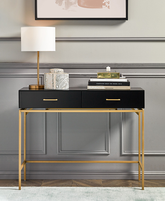 Flush up against a textured grey wall, the black and gold hallway console has a lamp and a stack of books styled on top.