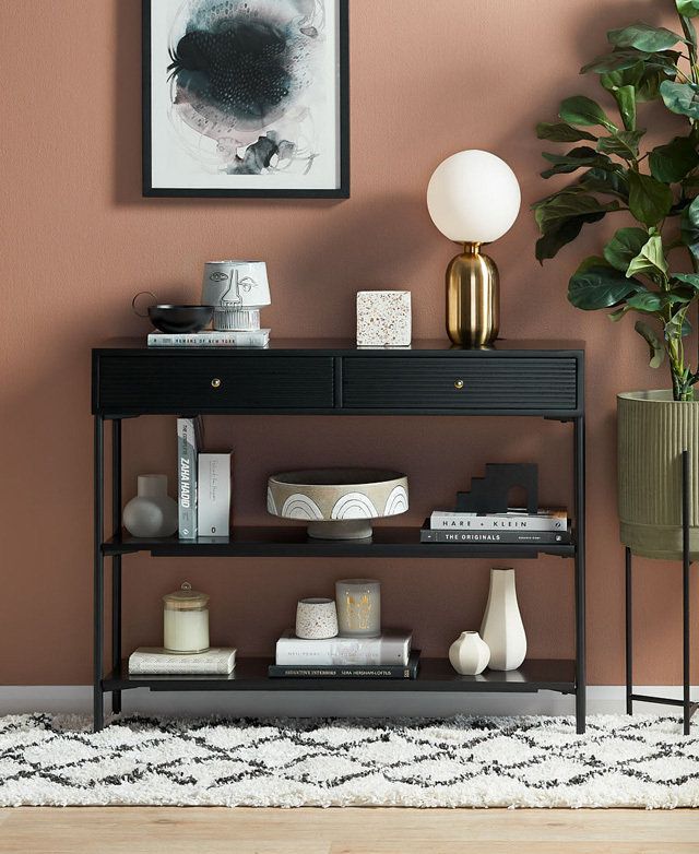 Myriad objects, books & a lamp pop against the black surfaces of the console, which is next to a peach wall & a plant.