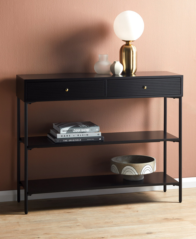 In front of a warm, peachy wall, the console is fully assembled and shown with a modern lamp, books, and vessels.
