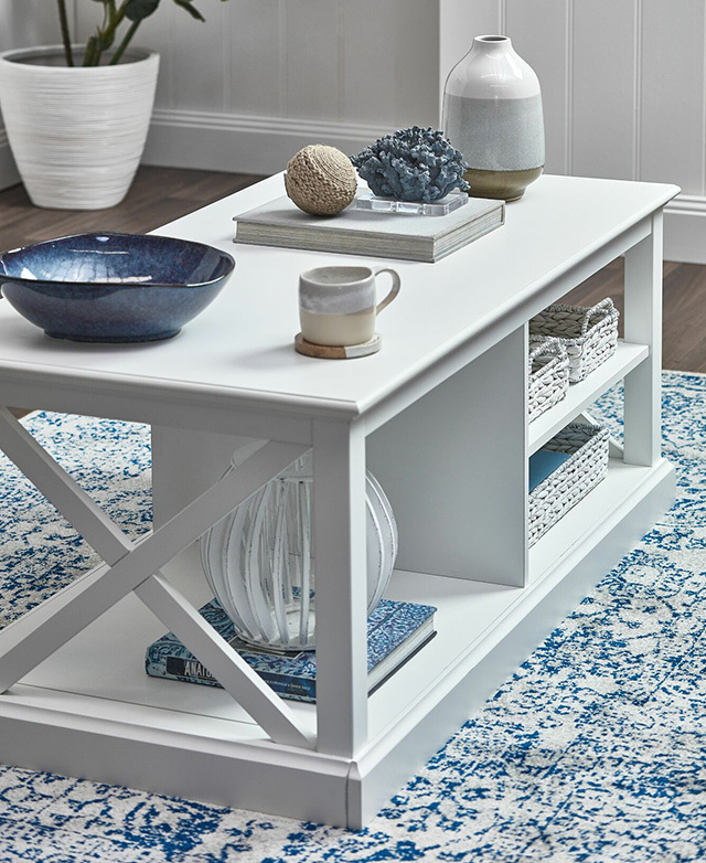 The white table is on a patterned blue rug in a bright room, with white and blue decor items on the surface areas. The white table is on a patterned blue rug in a bright room, with white and blue decor items on the surface areas.