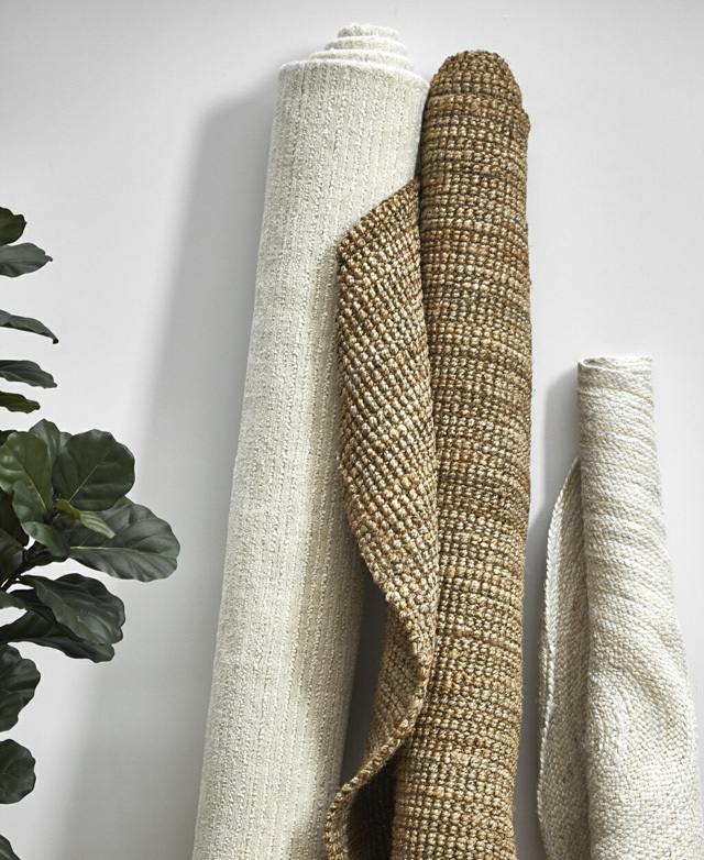 The rug is rolled and standing vertically against a wall next to two similarly rolled jute rugs in varying neutral hues.